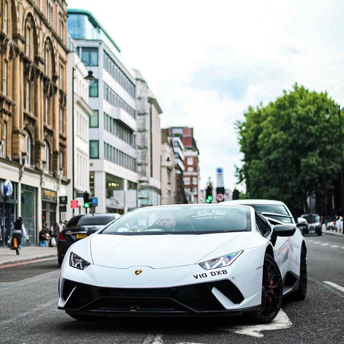 A white Lamborghini, available for hire, is driving down a city street surrounded by buildings and trees.