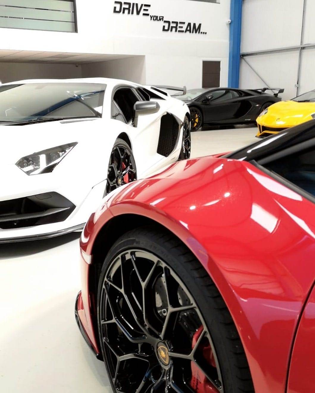 A showroom with four luxury sports cars in white, black, yellow, and red. A "DRIVE your DREAM..." sign is visible in the background, inviting you to hire one for an unforgettable experience.