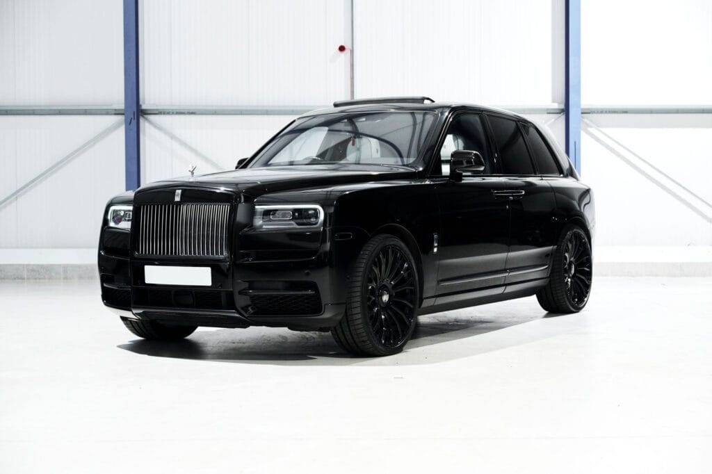 A sleek black luxury SUV with tinted windows is parked in a bright, minimalist indoor setting. Available for hire, the vehicle features a prominent grille and stylish black rims.