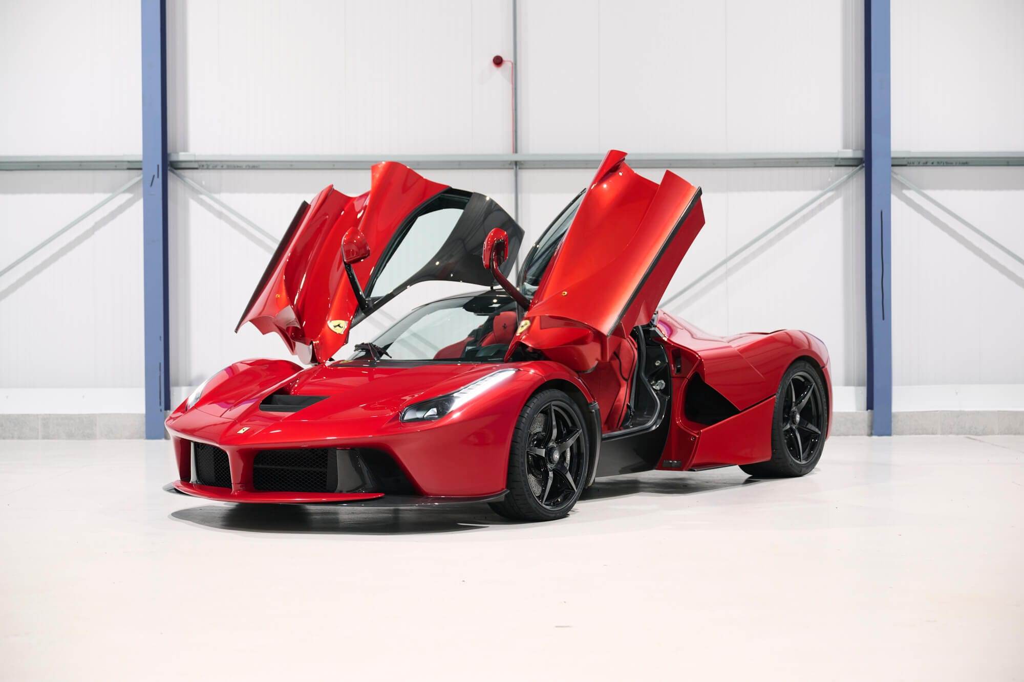 A red sports car with gull-wing doors open in a sleek, white indoor setting. The car features a stylish design and black wheels, ready to be hired for your next adventure.