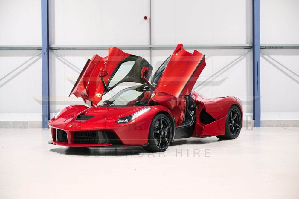 A red sports car with its scissor doors open is parked indoors in front of a white wall, available for hire. The car boasts black wheels and a low-profile design, offering both style and performance.