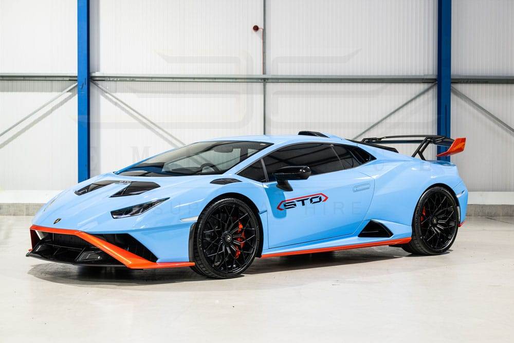 A light blue Lamborghini Huracán STO with orange and black accents is parked in a garage, flaunting its prominent rear wing and black wheels. This stunning supercar is available for hire, promising an unforgettable driving experience.