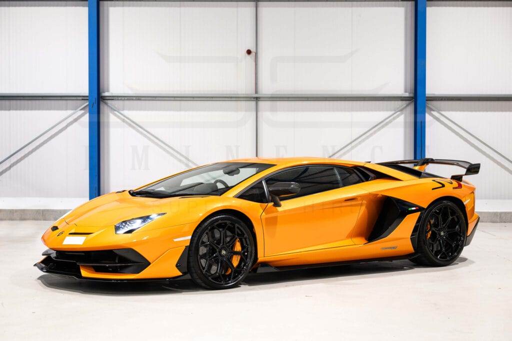 A yellow sports car with black accents is parked indoors. It has sleek, aerodynamic lines and a prominent rear spoiler, making it an attractive option to hire for your next adventure.