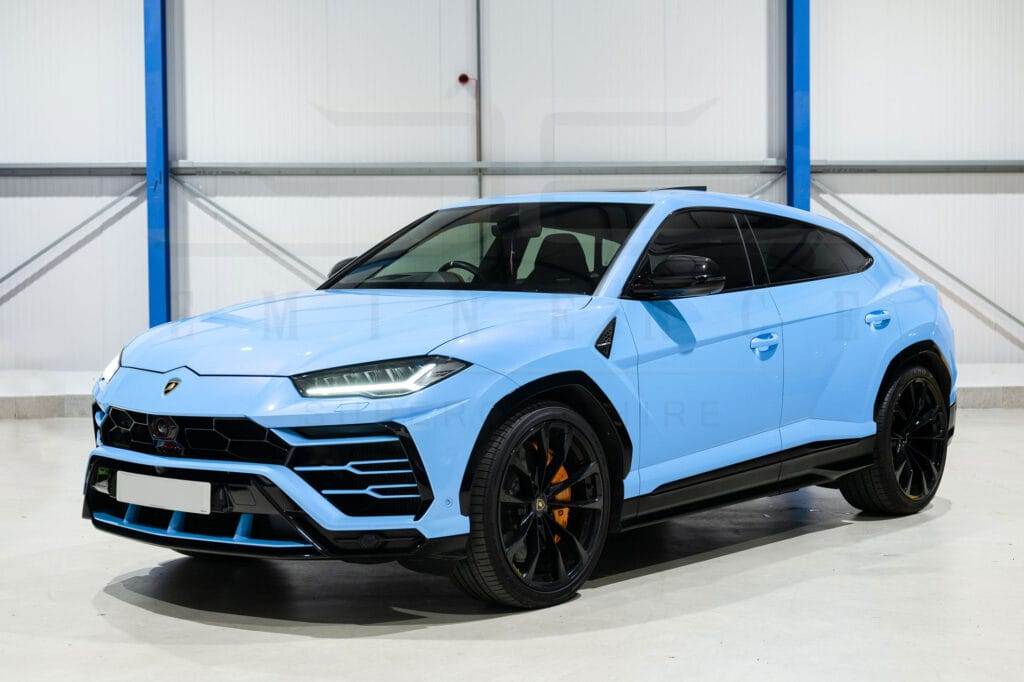 A light blue Lamborghini SUV, available for hire, is parked indoors in a well-lit garage with a sleek white and blue color scheme.