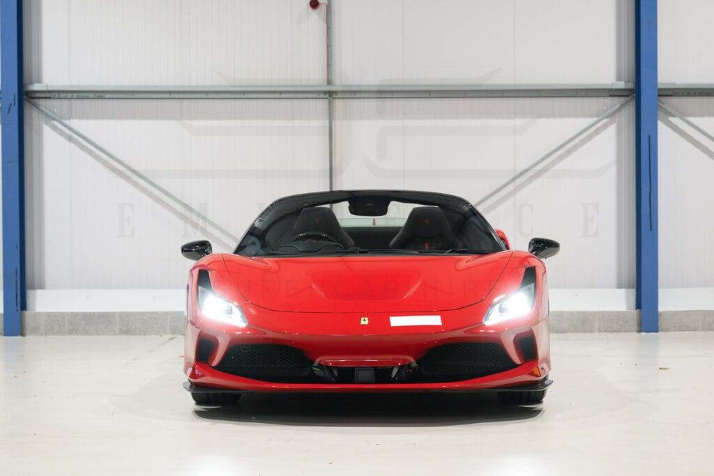 A red convertible sports car with headlights on, shown from the front, is parked indoors against a white and blue wall, ready for hire.