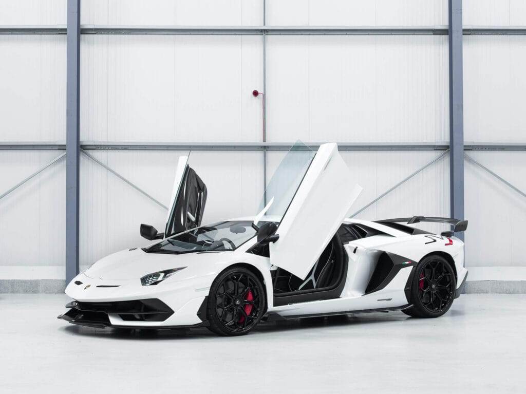 A sleek white sports car with both scissor doors open, parked in a modern garage. The car has black wheels with red brake calipers, ready for you to hire and hit the road in style.