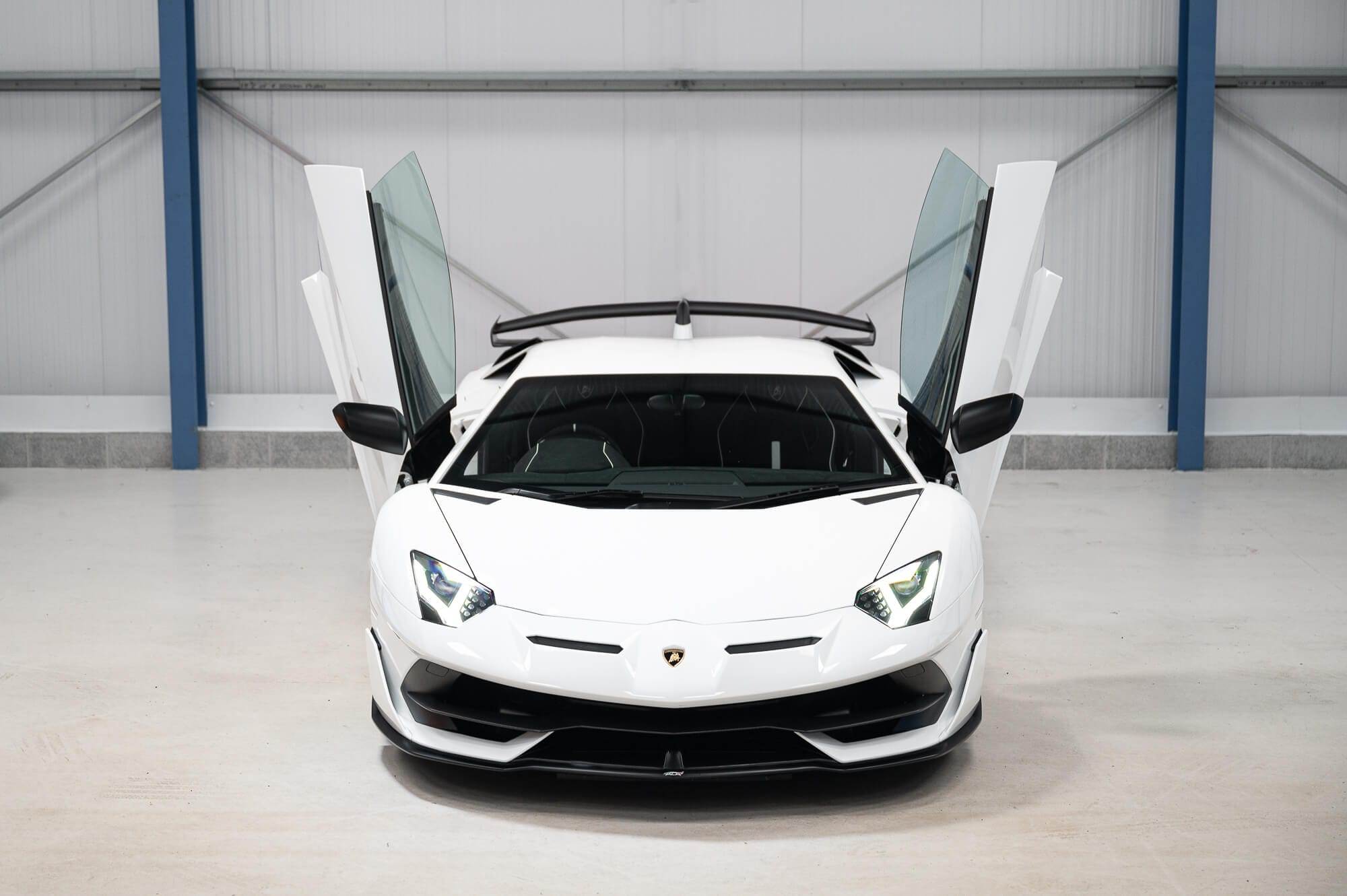 A white sports car with scissor doors open, shown from the front in a garage setting, available for hire.