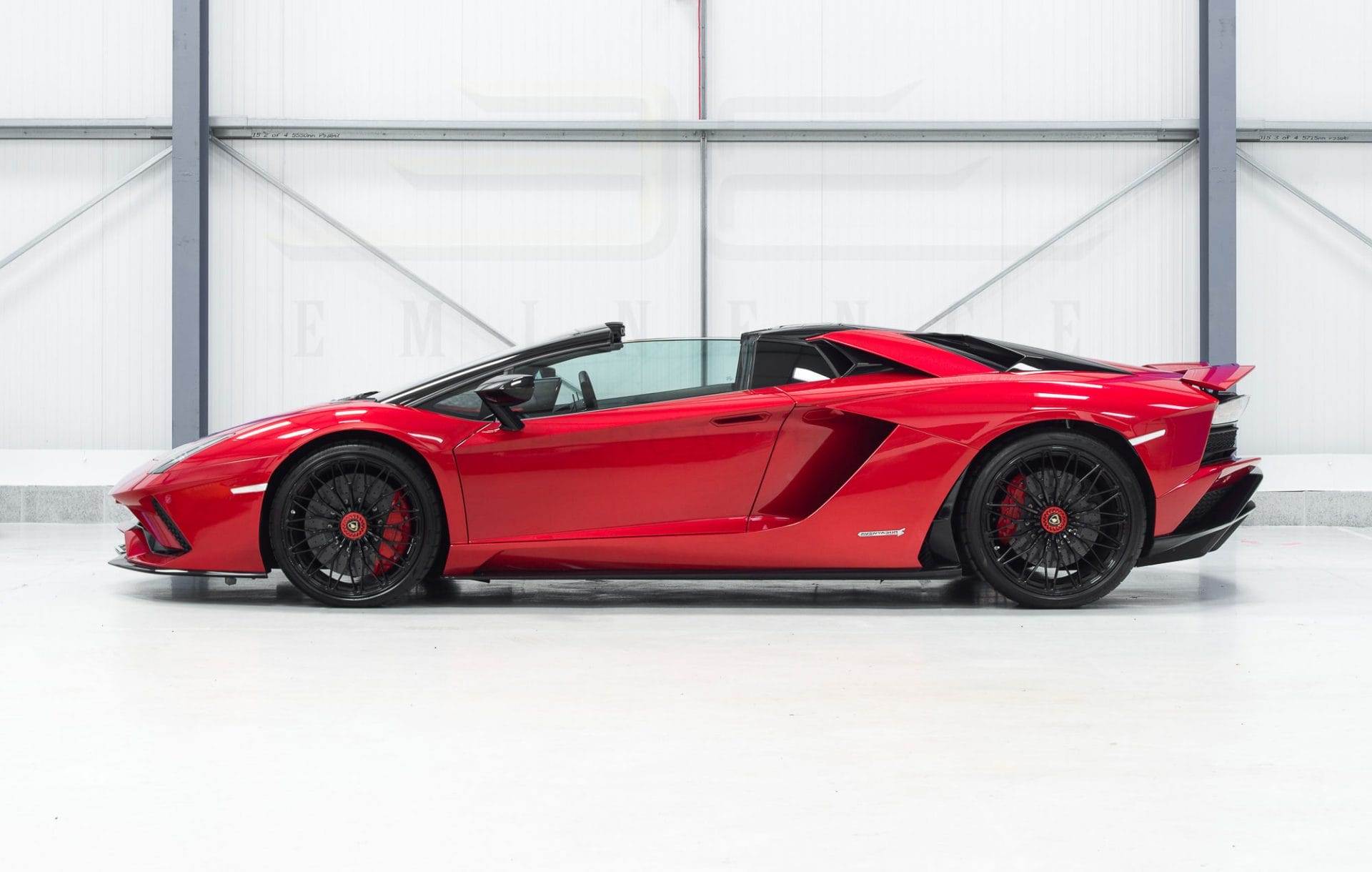 A sleek, red, high-performance sports car with a convertible top, parked indoors on a clean white floor, is available for hire.