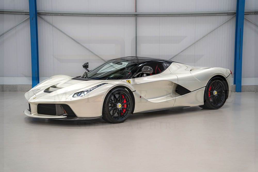 A sleek silver sports car with a black roof, parked indoors on a concrete floor. The car features an aerodynamic design, prominent side mirrors, and red brake calipers on black alloy wheels. Available for hire, this stunning vehicle is perfect for those seeking both style and performance.