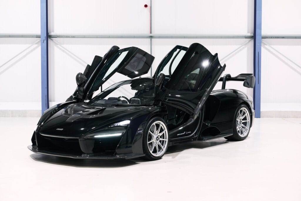 A black supercar with butterfly doors open, parked in an indoor garage, available for hire. The car features sleek aerodynamics, large front vents, and is positioned at a slight angle, showcasing both front and side views.