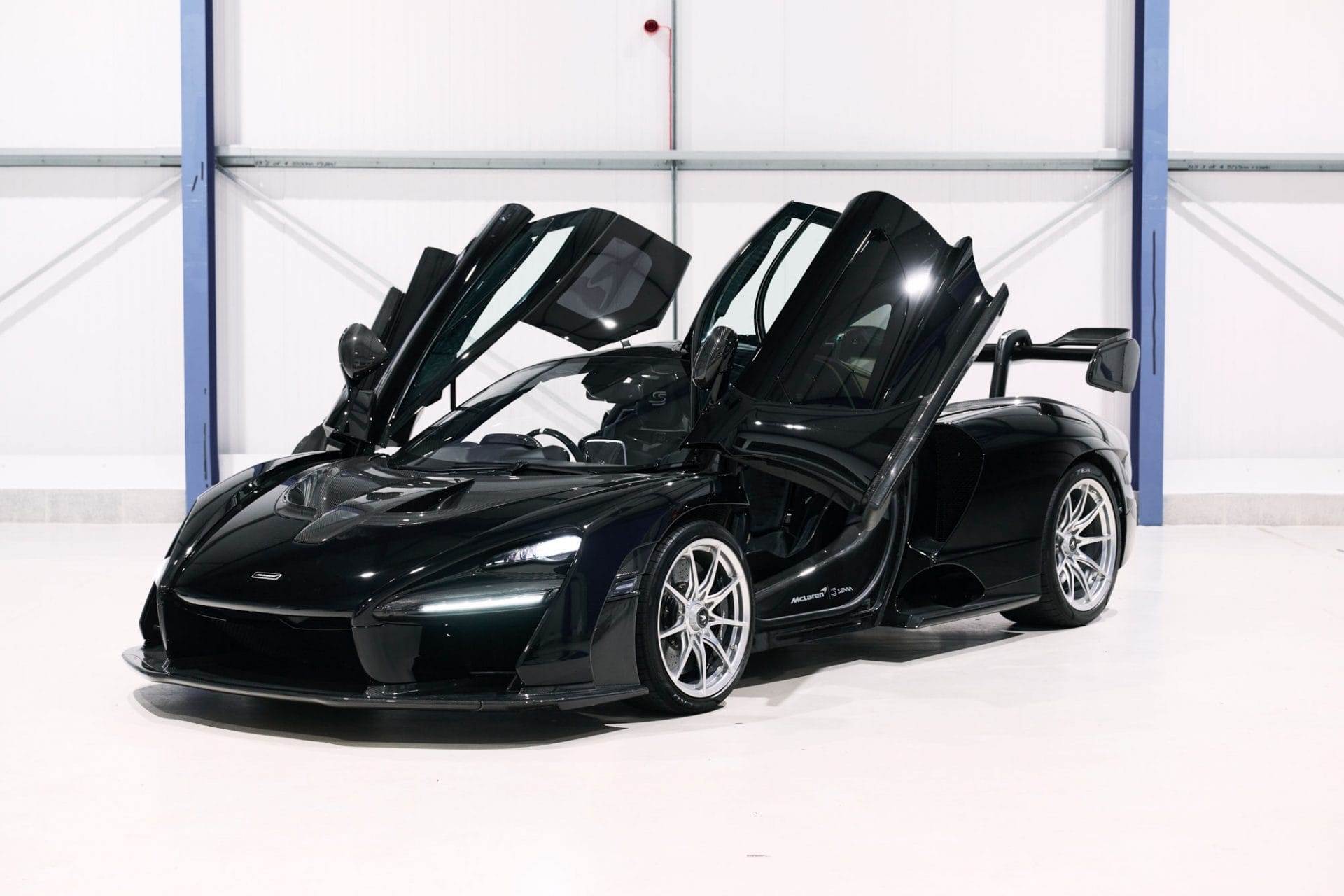 A sleek black sports car with scissor doors open, available for hire, parked in a minimalist white and blue indoor setting.