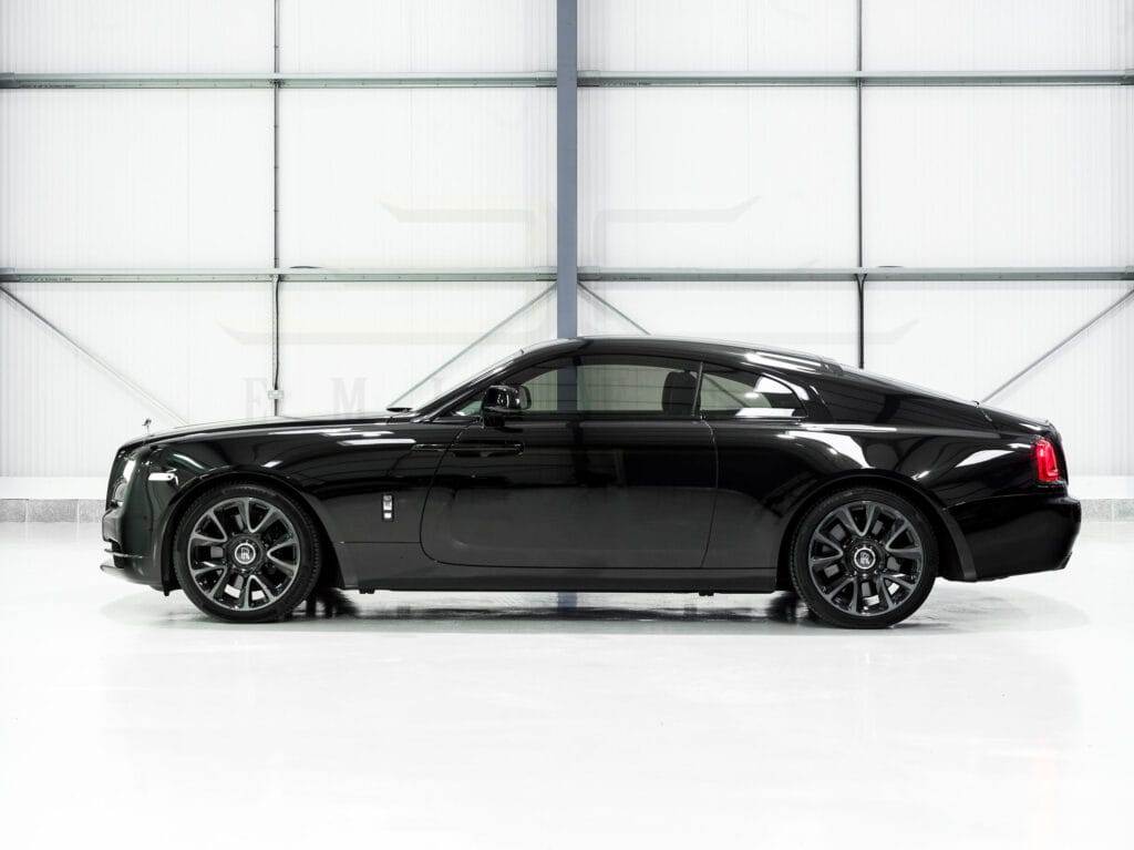 Side view of a sleek, black luxury coupe car parked in a spacious, well-lit indoor area with a reflective floor and industrial-style walls, available for hire.