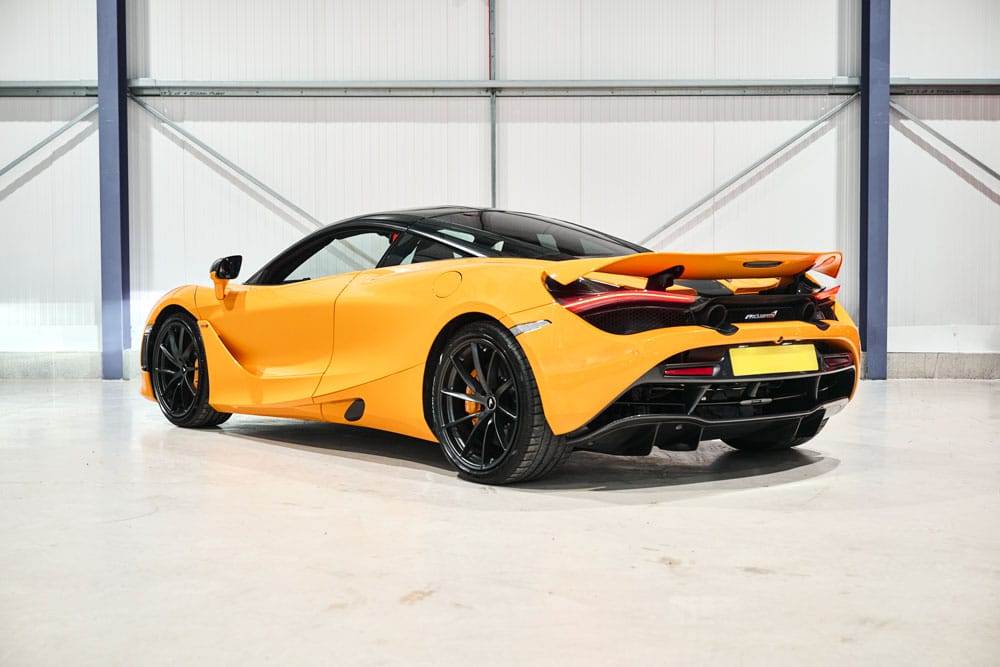 A bright orange sports car with black accents and a rear wing is parked inside a garage with a white floor and metal walls, ready for hire.