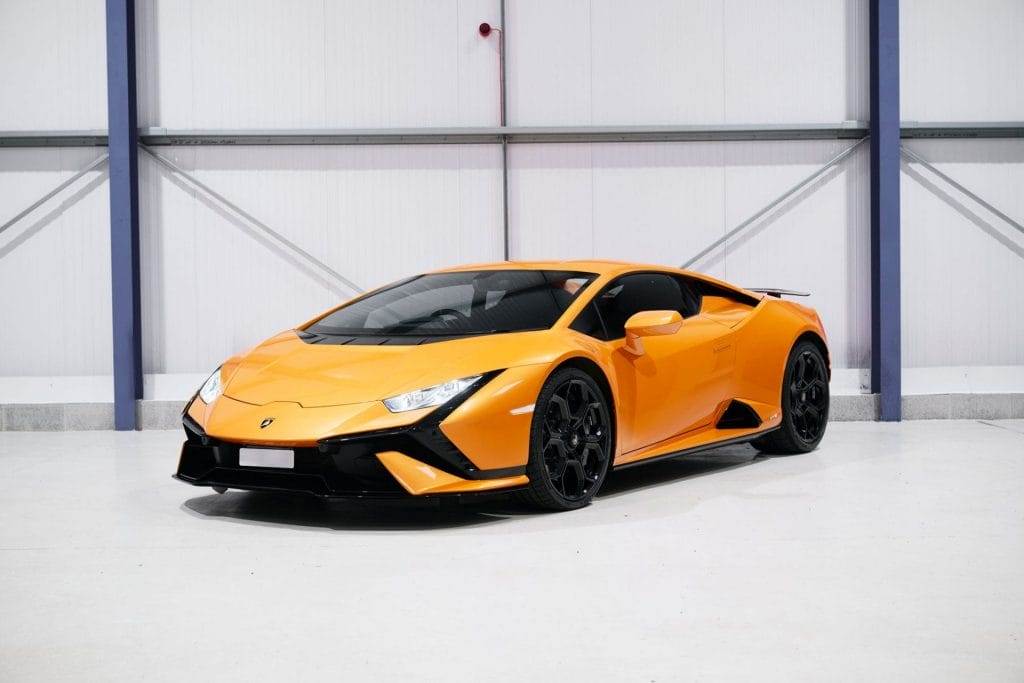 A bright orange sports car with black wheels, available for hire, parked indoors against a white and gray industrial background.